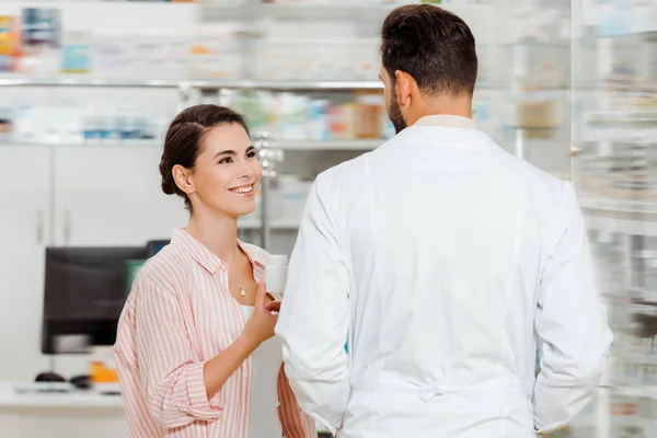 Un client souriant tenant un pot avec des pilules et regardant un pharmacien en pharmacie — Photo de stock