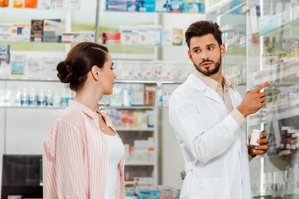 Druggist with pills pointing with finger at shelves with medicaments to customer — Stock Photo