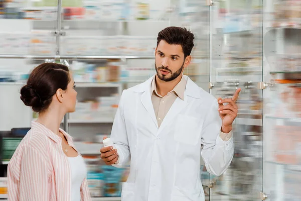 Druggist with pills pointing at medicaments in showcase to customer — Stock Photo