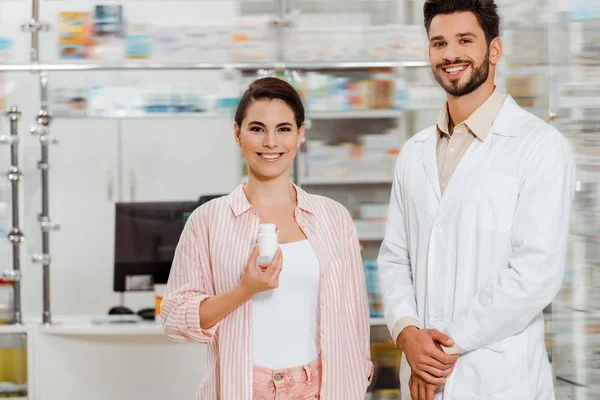 Mujer sonriente con frasco de píldoras al lado del farmacéutico con escaparate de farmacia en segundo plano - foto de stock