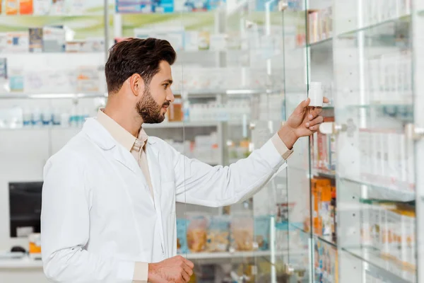 Side view of handsome pharmacist looking at jar with pills beside shelves with medicaments — Stock Photo