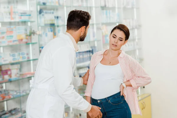 Druggist helping pregnant woman with pharmacy showcase at background — Stock Photo