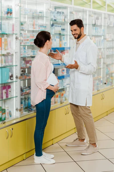 Lächelnder Apotheker schaut schwangere Kundin nach Medikamenten im Regal an — Stockfoto