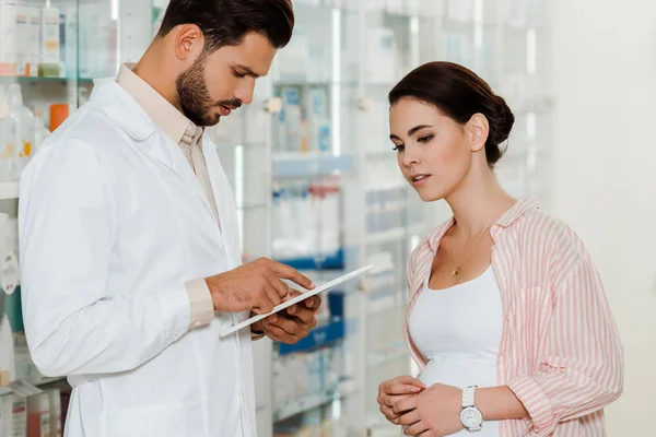 Druggist using digital tablet beside pregnant customer with medicaments on shelves at background — Stock Photo