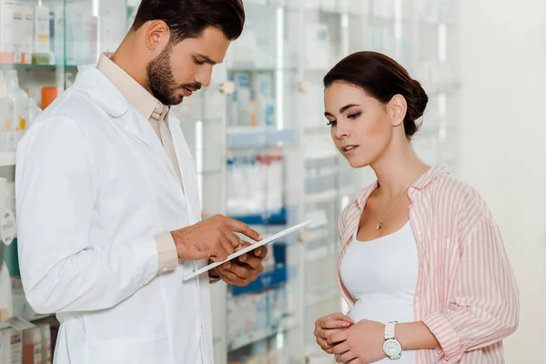 Druggist using digital tablet beside pregnant customer with medicaments on shelves at background — Stock Photo