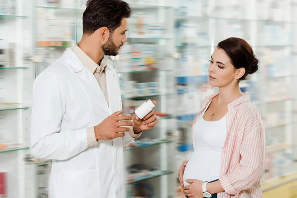 Vista lateral de la farmacia sosteniendo frasco con pastillas al lado de la mujer embarazada en boticario - foto de stock