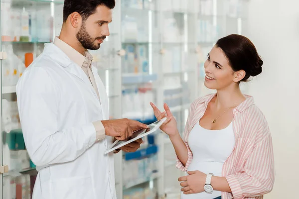 Vista lateral del farmacéutico con tableta digital mirando a la mujer embarazada sonriente en la farmacia - foto de stock