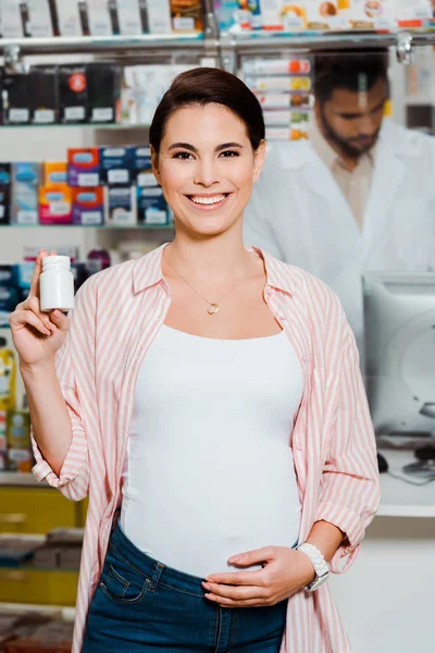 Femme enceinte souriant à la caméra et tenant pot avec des pilules avec pharmacien en arrière-plan — Photo de stock