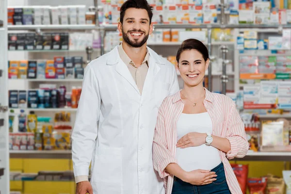 Улыбающийся фармацевт и беременная женщина смотрят в камеру с витриной аптеки на заднем плане — стоковое фото