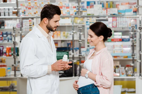 Side view of pharmacist showing jar with pills to pregnant smiling woman in apothecary — Stock Photo