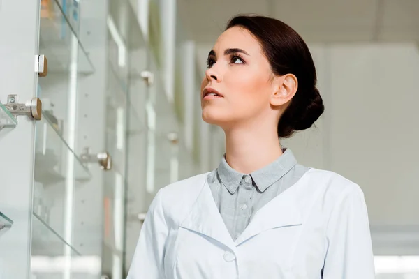 Low angle view of attractive pharmacist looking at medicaments in drugstore showcase — Stock Photo