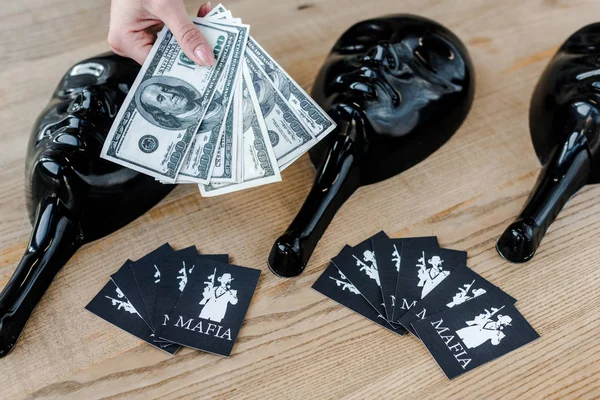 KYIV, UKRAINE - NOVEMBER 22, 2019: cropped view of woman holding dollar banknotes near playing cards with mafia letters and party masks on table — Stock Photo