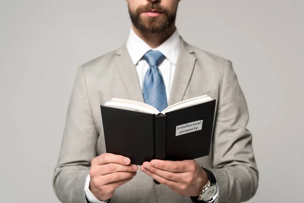 Vista recortada del hombre de negocios leyendo libro con título de propiedad intelectual aislado en gris - foto de stock