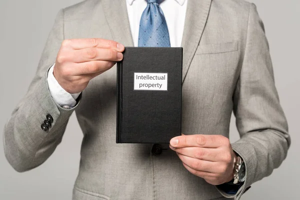Частичный взгляд бизнесмена, держащего юридическую книгу с названием интеллектуальной собственности изолированной на сером — стоковое фото