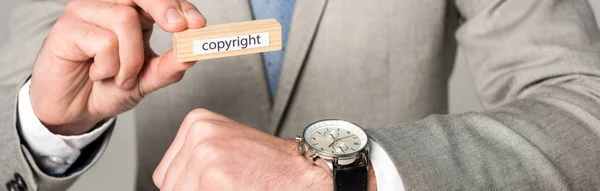 Vista recortada del hombre de negocios en reloj de pulsera que sostiene el bloque de madera con letras de derechos de autor aislados en gris, tiro panorámico - foto de stock