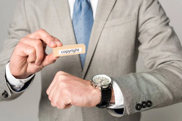 Vista recortada del hombre de negocios en reloj de pulsera que sostiene el bloque de madera con letras de derechos de autor aislados en gris - foto de stock