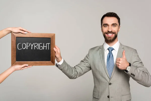 Vista recortada de la mujer sosteniendo pizarra con inscripción de derechos de autor cerca de hombre de negocios mostrando pulgar hacia arriba aislado en gris — Stock Photo