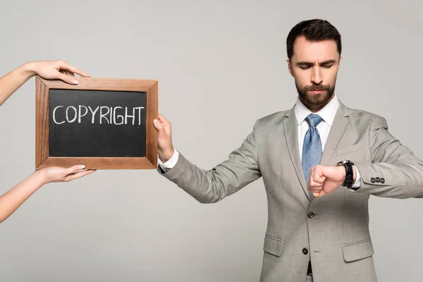 Vista recortada de la mujer sosteniendo pizarra con inscripción de derechos de autor cerca de hombre de negocios mirando reloj de pulsera aislado en gris - foto de stock