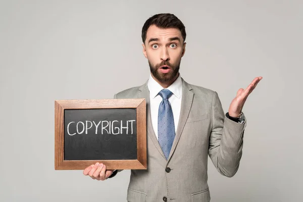 Shocked businessman looking at camera while holding chalkboard with copyright inscription isolated on grey — Stock Photo