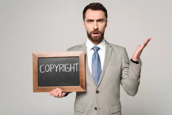Empresário descontente olhando para a câmera enquanto segurando quadro com inscrição de direitos autorais isolado em cinza — Fotografia de Stock