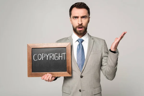 Displeased businessman looking at camera while holding chalkboard with copyright inscription isolated on grey — Stock Photo