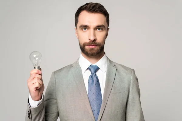 Confident businessman holding light bulb and looking at camera isolated on grey — Stock Photo
