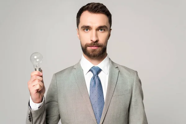 Hombre de negocios confiado sosteniendo la bombilla y mirando a la cámara aislada en gris — Stock Photo