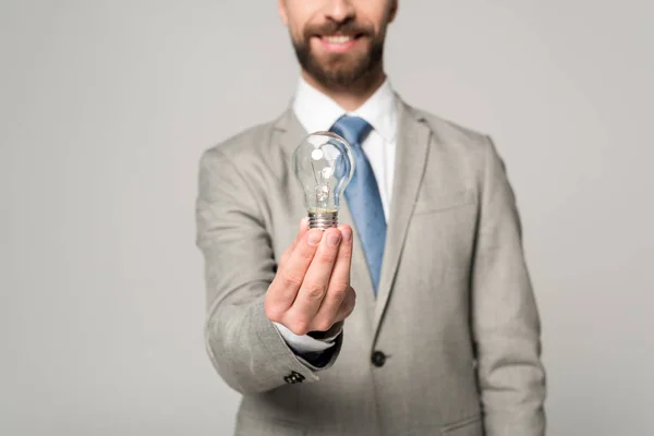 Vista parcial de un empresario sonriente sosteniendo una bombilla aislada en gris - foto de stock