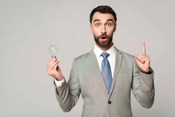 Excited businessman holding light bulb and showing idea sign isolated on grey — Stock Photo