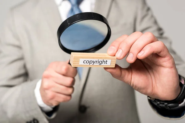 Cropped view of businessman looking at wooden block with word copyright through magnifier isolated on grey — Stock Photo