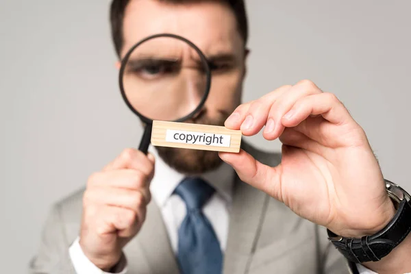 Enfoque selectivo de hombre de negocios mirando bloque de madera con derechos de autor palabra a través de lupa aislada en gris - foto de stock