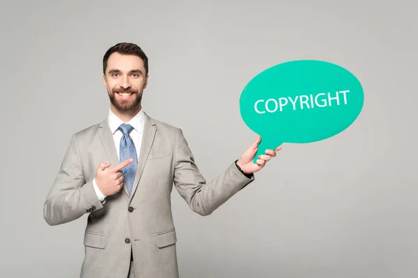 Uomo d'affari sorridente che punta con il dito alla bolla di pensiero con l'iscrizione di copyright isolata su grigio — Foto stock