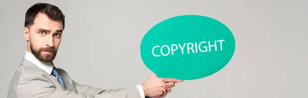 Panoramic shot of serious businessman holding thought bubble with copyright inscription isolated on grey — Stock Photo