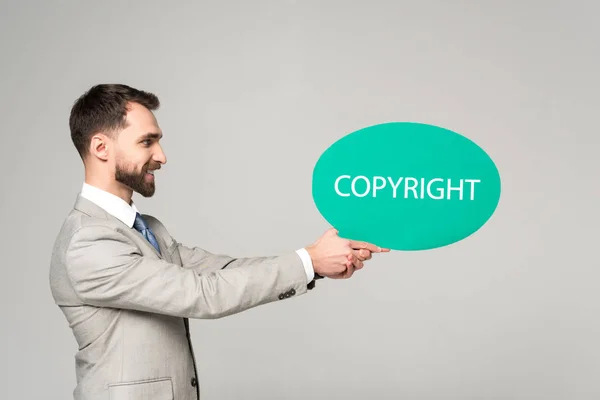 Vue latérale d'un homme d'affaires souriant tenant une bulle de pensée avec inscription de copyright isolée sur gris — Photo de stock