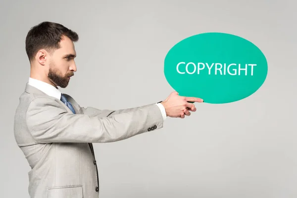 Vista lateral del empresario confiado sosteniendo la burbuja del discurso con el copyright de la palabra aislado en gris — Stock Photo