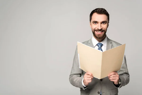Positiver Geschäftsmann lächelt in die Kamera, während er Papiermappe isoliert auf grau hält — Stockfoto