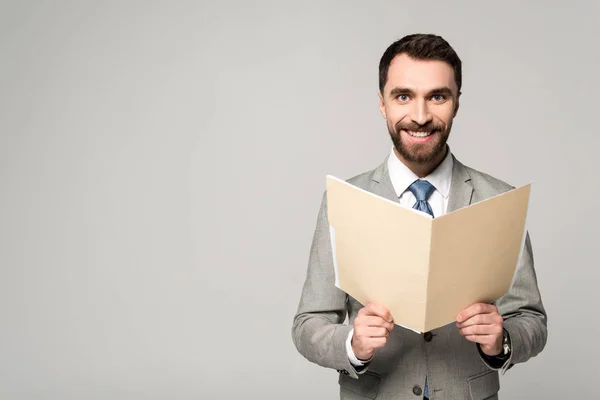 Hombre de negocios positivo sonriendo a la cámara mientras sostiene la carpeta de papel aislada en gris - foto de stock