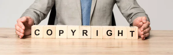 Partial view of businessman sitting at desk near wooden cubes with word copyright isolated on grey, panoramic shot — Stock Photo