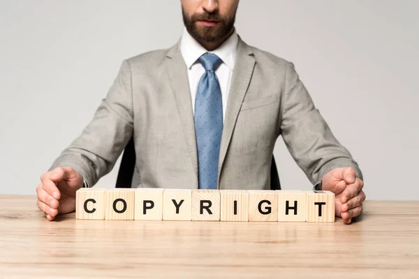 Vista recortada del hombre de negocios sentado en el escritorio cerca de cubos de madera con la palabra copyright aislado en gris - foto de stock