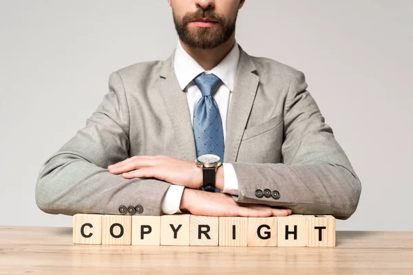 Vista parcial del hombre de negocios sentado en el escritorio cerca de cubos de madera con la palabra copyright aislado en gris - foto de stock