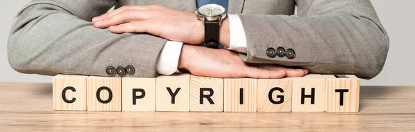 Partial view of businessman sitting at desk near wooden cubes with word copyright isolated on grey, panoramic shot — Stock Photo