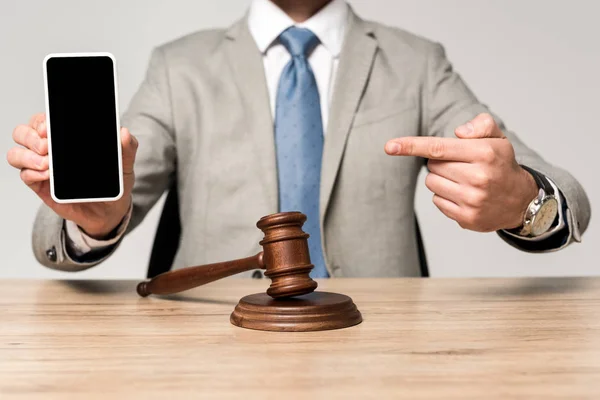 Vista ritagliata di avvocato che punta con il dito a smartphone con schermo bianco, e martelletto sulla scrivania isolato su grigio — Foto stock