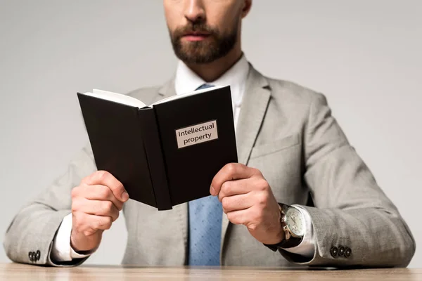 Vista recortada del hombre de negocios leyendo libro jurídico con título de propiedad intelectual aislado en gris - foto de stock