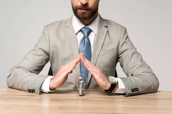Vista parcial del empresario mostrando gesto de protección por encima de la bombilla aislada en gris - foto de stock