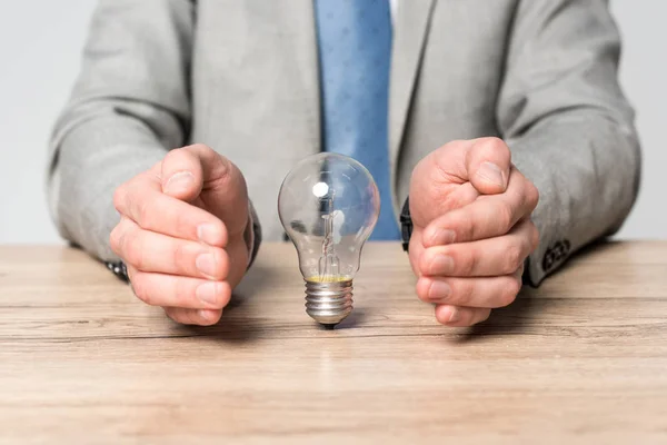 Partial view of businessman showing protection gesture near light bulb isolated on grey — Stock Photo