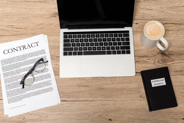 Vue du dessus de l'ordinateur portable, contrat, lunettes, tasse de café et livre de propriété intellectuelle sur bureau en bois — Photo de stock