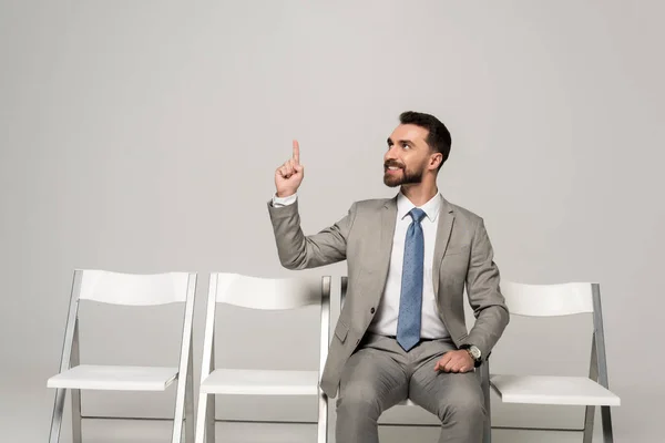 Усміхнений бізнесмен сидить на стільці і показує жест ідеї ізольований на сірому — стокове фото