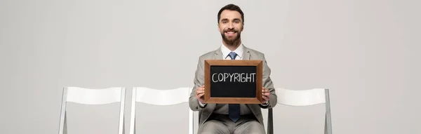Plano panorámico de empresario feliz sosteniendo pizarra con derechos de autor palabra mientras está sentado en silla aislada en gris - foto de stock