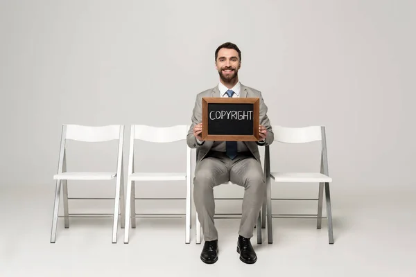 Homme d'affaires heureux tenant tableau noir avec droit d'auteur mot tout en étant assis sur la chaise sur fond gris — Photo de stock