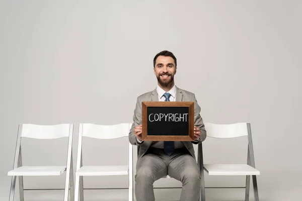 Smiling businessman holding chalkboard with word copyright while sitting on chair on grey background — Stock Photo